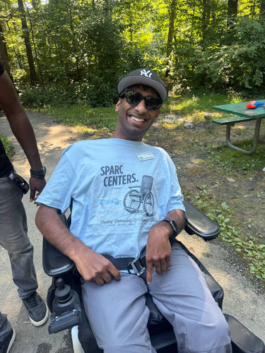 Smiling SPARC participant in a power wheelchair wearing a SPARC Centers t-shirt, enjoying a sunny day outdoors at a park.